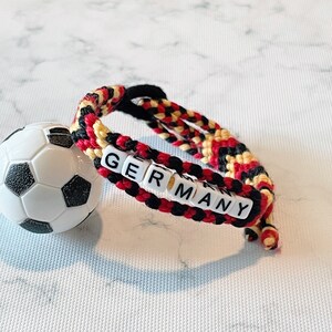 K&ouml;nnte beinhalten: Ein geflochtenes Armband in den Farben der deutschen Flagge: Schwarz, Rot und Gelb. Das Wort "GERMANY" ist mit wei&szlig;en Buchstabenperlen geschrieben. Ein kleiner Fu&szlig;ball liegt neben dem Armband.