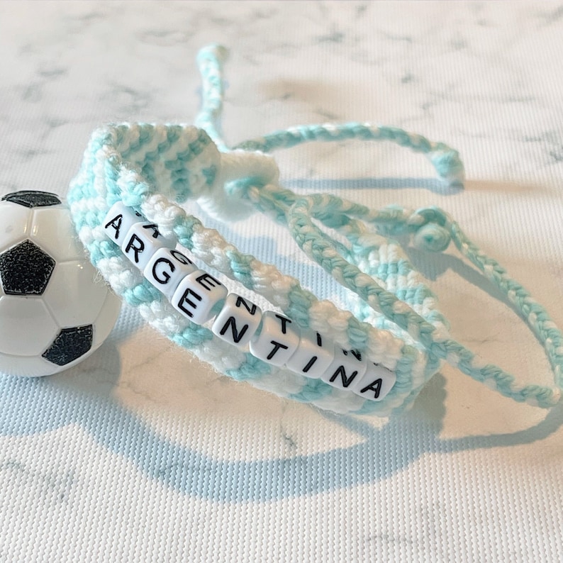 K&ouml;nnte beinhalten: Handgefertigtes Armband mit dem Wort "ARGENTINA" aus wei&szlig;en Buchstabenperlen. Das Armband ist aus hellblauer und wei&szlig;er Wolle gefertigt und liegt neben einem kleinen Fu&szlig;ball.