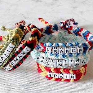 K&ouml;nnte beinhalten: Handgefertigte H&auml;kelarmb&auml;nder in verschiedenen Farben, mit L&auml;ndernamen, die in wei&szlig;en Buchstabenperlen geschrieben sind. Die Armb&auml;nder tragen die Namen "Brasilien", "Deutschland", "Frankreich", "Argentinien" und "Spanien". Die Armb&auml;nder sind gestapelt.