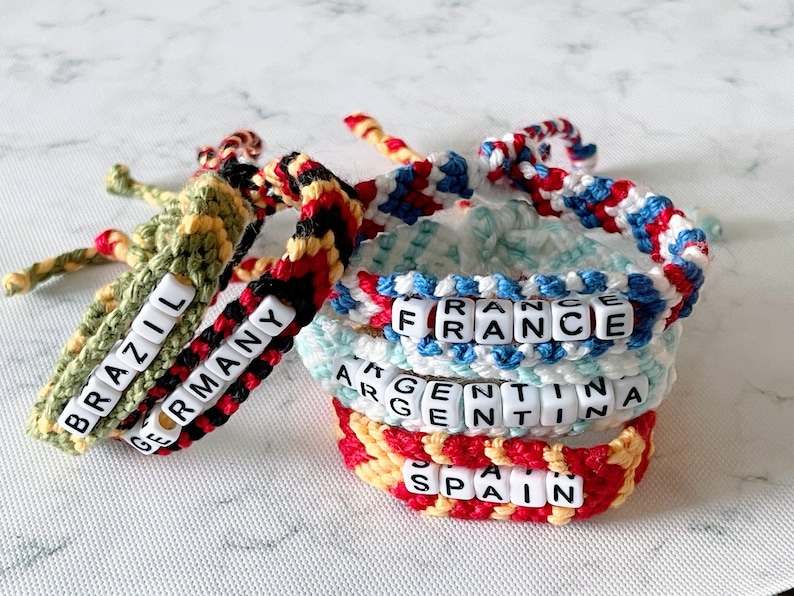 K&ouml;nnte beinhalten: Handgefertigte H&auml;kelarmb&auml;nder in verschiedenen Farben, mit L&auml;ndernamen, die in wei&szlig;en Buchstabenperlen geschrieben sind. Die Armb&auml;nder tragen die Namen "Brasilien", "Deutschland", "Frankreich", "Argentinien" und "Spanien". Die Armb&auml;nder sind gestapelt.