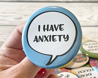 May include: A round, light blue button with the text "I HAVE ANXIETY" in a white speech bubble. The button is held in a hand with other buttons visible in the background.