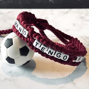 K&ouml;nnte beinhalten: Ein burgunderfarbenes Makramee-Armband mit wei&szlig;en Buchstabenperlen mit der Aufschrift "MANO TENGO FE". Das Armband liegt neben einem kleinen Fu&szlig;ball. Es ist ein handgefertigter Artikel.