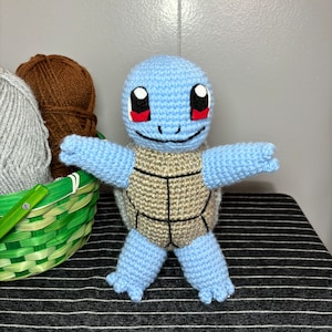 May include: A blue and tan crocheted Squirtle plush toy with red eyes. The toy is standing on a striped surface.