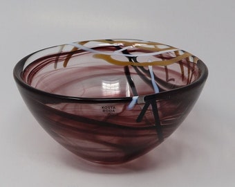 May include: A small, clear glass bowl with a swirling pattern of purple, black, white, and gold. The bowl is made by Kosta Boda.