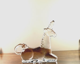 May include: A clear glass unicorn figurine with a brown body and white mane and horn. The unicorn is in a seated position with its head held high. The figurine is on a wooden surface.