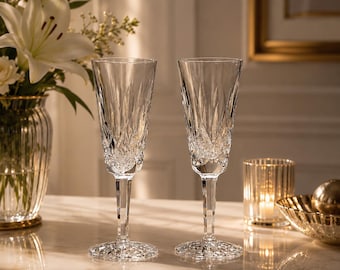 May include: Two clear crystal champagne flutes featuring a vertical diamond-cut pattern on the bowl and a faceted stem. The base of each glass has a radial starburst design, reflecting light across the surface with a polished, transparent finish.