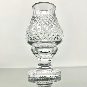 May include: A clear glass candle holder with a diamond-cut pattern on the bowl-shaped top. The holder has a pedestal base and a flared rim. The glass reflects light, creating a sparkling effect. The candle holder is designed to hold a small candle.
