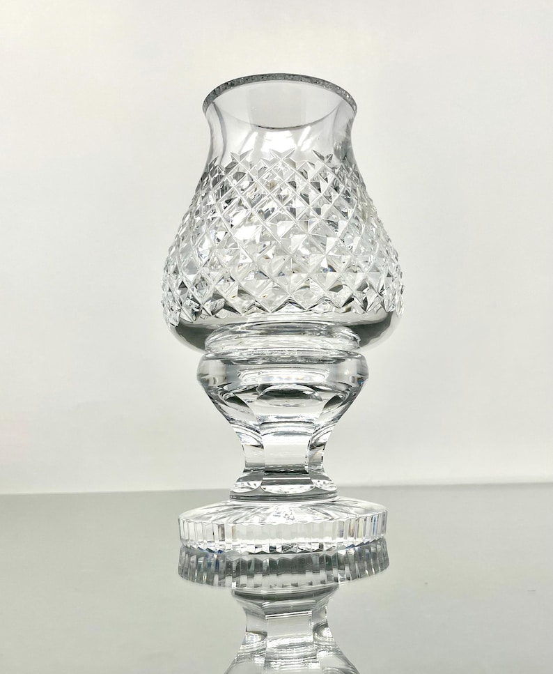 May include: A clear glass candle holder with a diamond-cut pattern on the bowl-shaped top. The holder has a pedestal base and a flared rim. The glass reflects light, creating a sparkling effect. The candle holder is designed to hold a small candle.