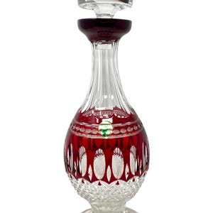 Puede incluir: Una decantador de vidrio transparente con un diseño de cristal rojo y un tapón de vidrio transparente. La decantador tiene un cuerpo redondo y un cuello alto y delgado. El tapón tiene forma de cúpula.