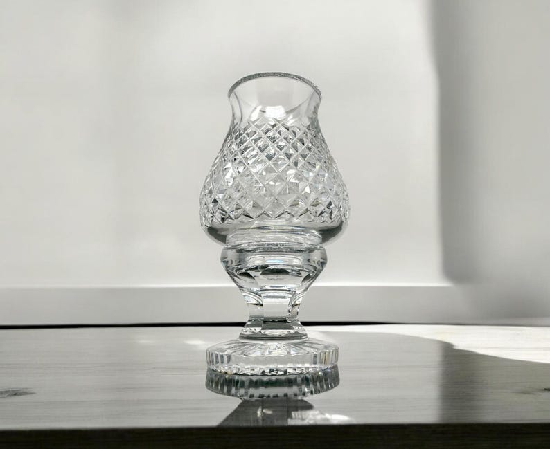 May include: A clear, cut-glass candle holder with a diamond pattern. The candle holder has a flared top, a bulbous midsection, and a pedestal base. The glass reflects light, creating a sparkling effect. The candle holder is designed to hold a small candle.