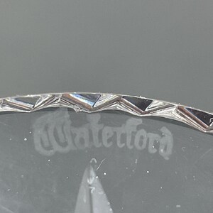 May include: Close-up of a clear crystal glass with the etched word "Waterford". The rim has a decorative, geometric design. The glass is set against a gray background.
