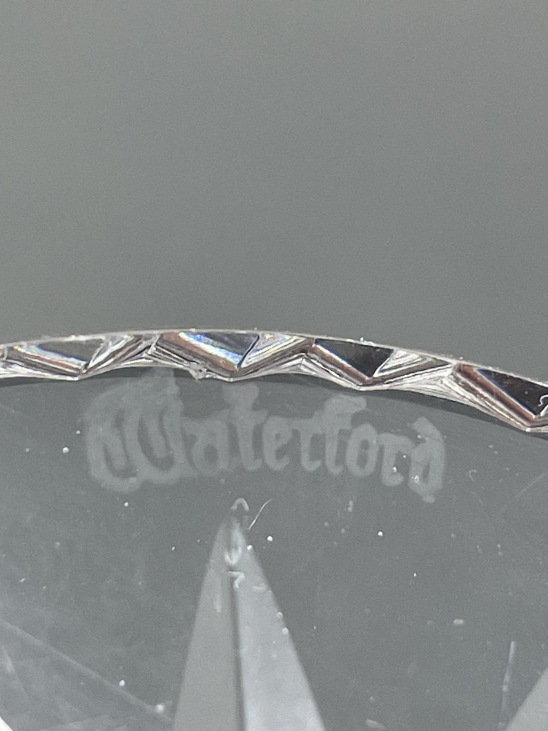 May include: Close-up of a clear crystal glass with the etched word "Waterford". The rim has a decorative, geometric design. The glass is set against a gray background.