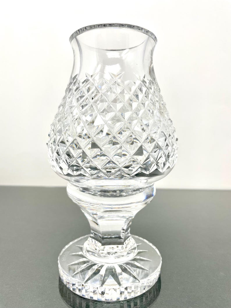 May include: A clear glass candle holder with a diamond-cut pattern on the body. The base is round with a starburst design. The candle holder has a flared top and a pedestal base.