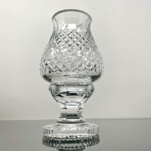 May include: Clear glass candle holder with a diamond-cut pattern. The candle holder has a flared top, a bulbous midsection, and a pedestal base. The glass reflects light, creating a sparkling effect. The overall design is elegant and classic.