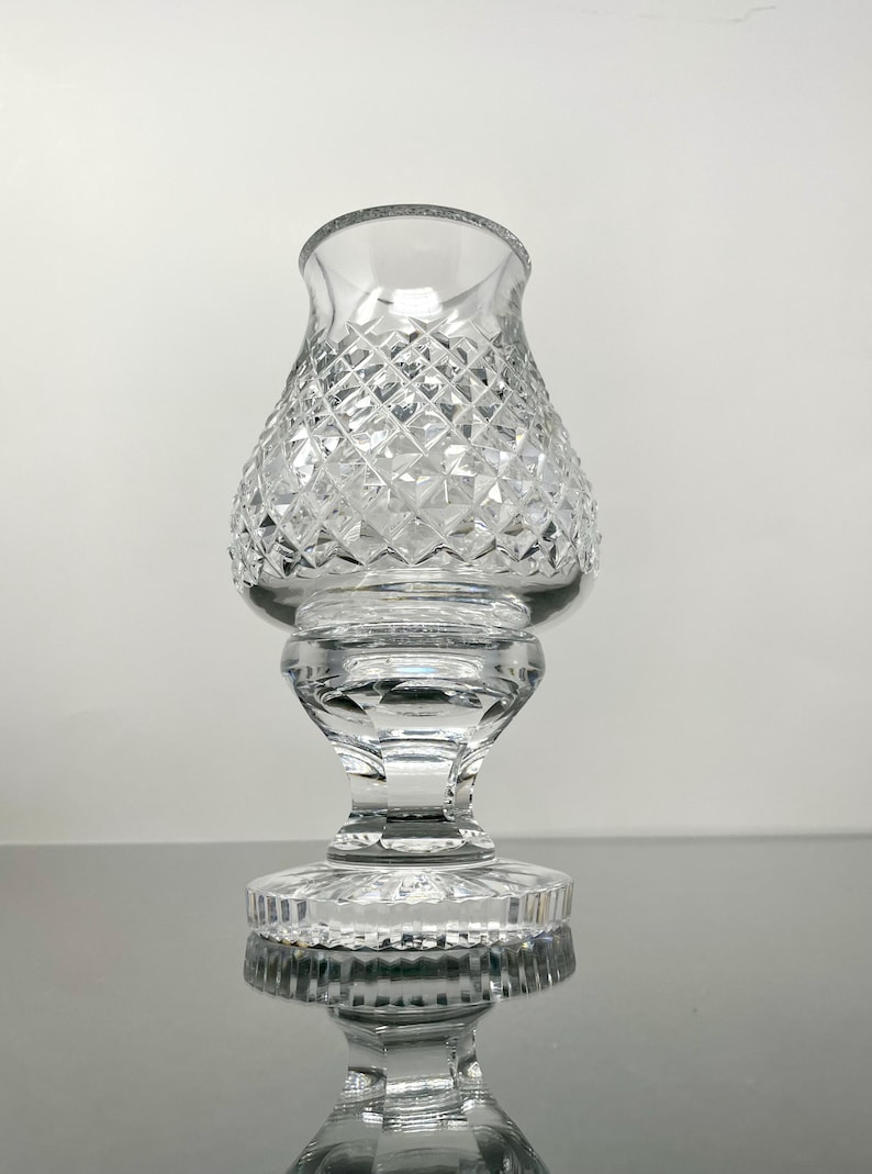 May include: Clear glass candle holder with a diamond-cut pattern. The candle holder has a flared top, a bulbous midsection, and a pedestal base. The glass reflects light, creating a sparkling effect. The overall design is elegant and classic.