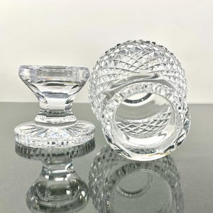 May include: Two clear, cut-glass items. One is a small, round, diamond-patterned glass with a wide opening. The other is a small, pedestal-style glass with a flat, round base and a flared top. Both items are highly reflective.