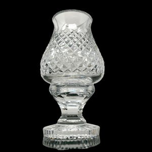 May include: A clear glass candle holder with a diamond-cut pattern. The candle holder has a wide base, a pedestal, and a flared top. The glass is transparent, allowing light to pass through. The candle holder is approximately 7 inches tall.
