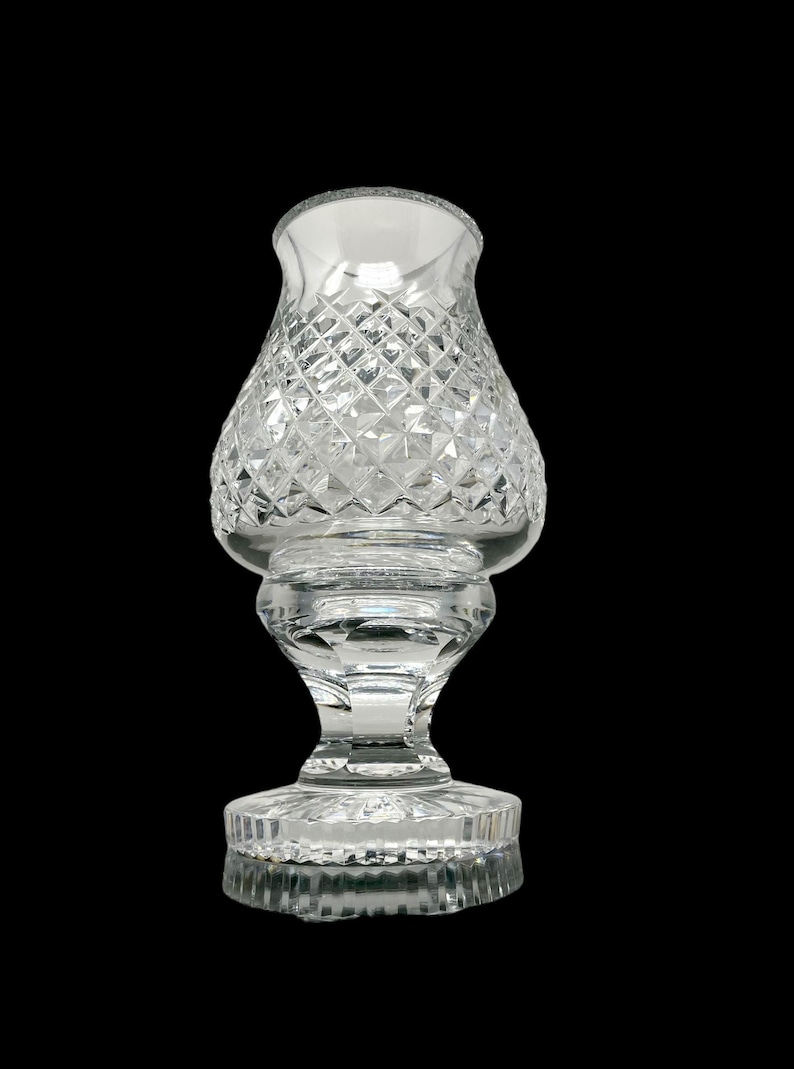 May include: A clear glass candle holder with a diamond-cut pattern. The candle holder has a wide base, a pedestal, and a flared top. The glass is transparent, allowing light to pass through. The candle holder is approximately 7 inches tall.