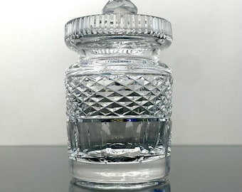May include: A clear, cut-glass jar with a lid. The jar has a diamond pattern on the body and vertical lines on the lid and top rim. The jar is sitting on a reflective surface.