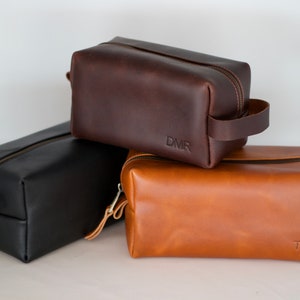 May include: Three leather toiletry bags in brown, black, and tan. The bags have zipper closures and leather handles. The brown bag has the initials "DMR" embossed on the front, the tan bag has the initials "TK" embossed on the front, and the black bag has no initials.
