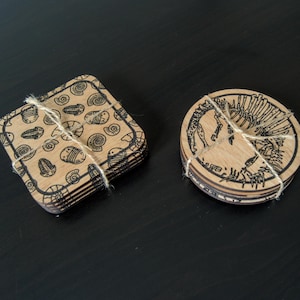K&ouml;nnte beinhalten: Satz mit sechs Untersetzern mit einem schwarz-wei&szlig;en Design. Ein Untersetzer zeigt ein Muster aus Muscheln und der andere eine stilisierte menschliche Figur. Die Untersetzer sind aus Holz und mit Schnur zusammengebunden.