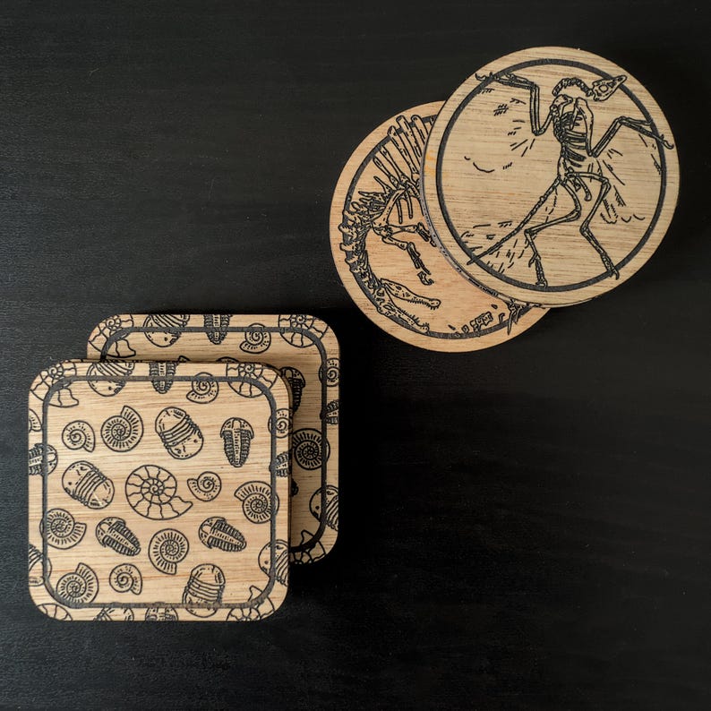 K&ouml;nnte beinhalten: Satz mit vier Untersetzern aus Holz mit verschiedenen Designs. Ein Untersetzer zeigt eine Schwarzwei&szlig;illustration eines Dinosaurierskeletts, w&auml;hrend die anderen drei Untersetzer eine Schwarzwei&szlig;illustration verschiedener versteinerter Meeresbewohner zeigen.