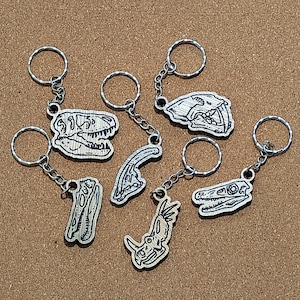 May include: A set of seven keychains featuring different dinosaur skeletons. Each keychain is made of wood and has a silver metal ring. The skeletons are all black and white and are drawn in a detailed style.
