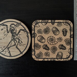 K&ouml;nnte beinhalten: Zwei Untersetzer aus Holz mit lasergravierten Designs. Ein Untersetzer zeigt ein schwarz-wei&szlig;es Skelett eines Vogels mit ausgebreiteten Fl&uuml;geln. Der andere Untersetzer hat ein sich wiederholendes Muster aus schwarzen und wei&szlig;en Trilobiten und Nautilus-Schalen.