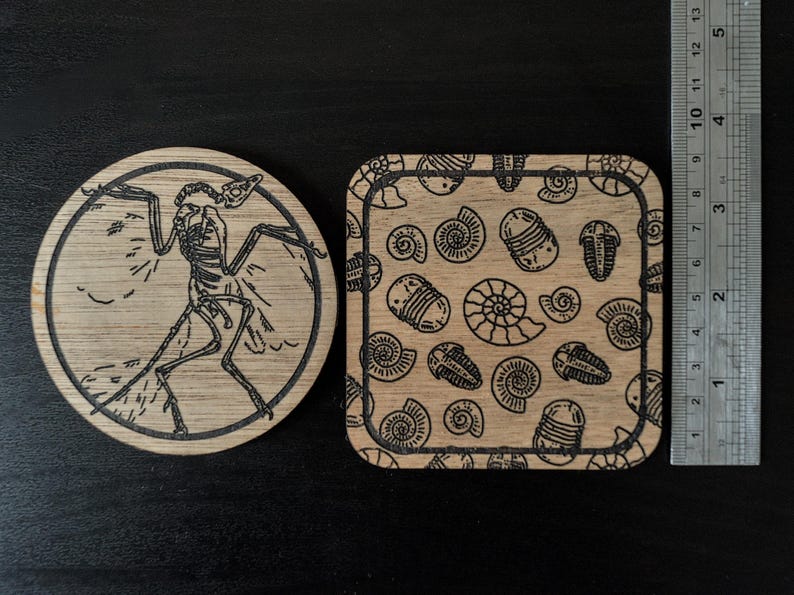 K&ouml;nnte beinhalten: Zwei Untersetzer aus Holz mit lasergravierten Designs. Ein Untersetzer zeigt ein schwarz-wei&szlig;es Skelett eines Vogels mit ausgebreiteten Fl&uuml;geln. Der andere Untersetzer hat ein sich wiederholendes Muster aus schwarzen und wei&szlig;en Trilobiten und Nautilus-Schalen.