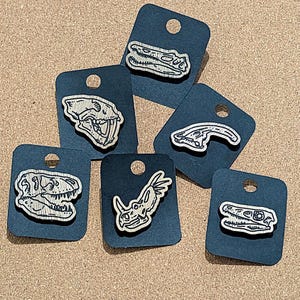 May include: Six black cardstock tags with white dinosaur skull illustrations. The tags are arranged in a semi-circle on a brown surface. The dinosaur skulls are of different species, including a T-Rex, Triceratops, and Stegosaurus.
