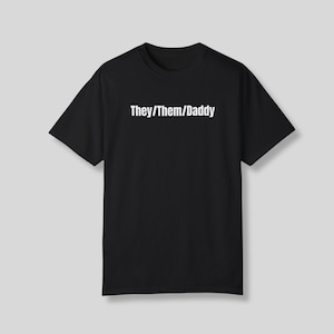 May include: Black t-shirt with the text "They/Them/Daddy" in white, centered on the chest. The shirt has short sleeves and a crew neck. The background is a solid light gray.
