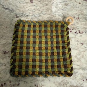 May include: A woven potholder in shades of green, brown, and grey, featuring a checkered pattern. The potholder has a braided edge and a wooden ring for hanging. This kitchen accessory is designed for handling hot cookware.