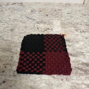 May include: A square, woven potholder in black and burgundy. The potholder is divided into two sections, with a checkerboard pattern in each. A small wooden ring is attached to one corner for hanging.