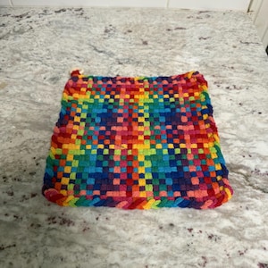 May include: A square, woven hot pad in a vibrant rainbow pattern. The pad features a tight weave of red, orange, yellow, green, blue, and purple threads. The edges are finished with a braided border in coordinating colours. This kitchen accessory is designed for heat resistance.