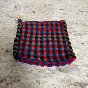 May include: A square, woven hot pad with a wooden ring for hanging. The pad features a checkerboard pattern of red, blue, black, and grey yarn. The edges are trimmed with a braided blue and red border.
