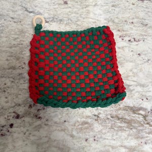 May include: A square, woven potholder in a red and green checkerboard pattern. The edges are trimmed with a thick, red border and green trim. A small, round wooden ring is attached to one corner for hanging.