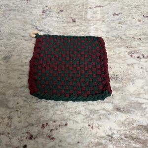 May include: A square, woven potholder in a checkerboard pattern of dark red and green. The edges are trimmed with a green border, and a small wooden ring is attached for hanging. This kitchen accessory is designed for heat resistance.