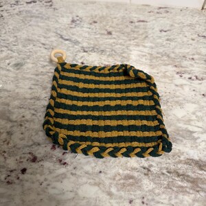 Op de afbeelding: Een vierkante, handgemaakte pannenlap met afwisselende horizontale strepen in donkergroen en goud. De randen zijn afgewerkt met een gevlochten donkergroene rand. Er is een kleine houten ring bevestigd om op te hangen.