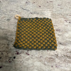 May include: A square, woven potholder in a checkerboard pattern of olive green and mustard yellow. It has a small, wooden ring attached to one corner for hanging. The potholder is designed for kitchen use.