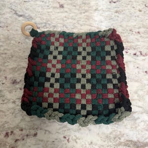 May include: Woven square hot pad in a checkerboard pattern of red, green, black, and grey. The edges are braided in a matching colour scheme. A small wooden ring is attached for hanging.