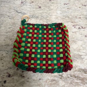 May include: A woven potholder with a checkerboard pattern of red, green, and burgundy squares. The edges are trimmed with a green and burgundy rope-like border. A small wooden ring is attached for hanging.