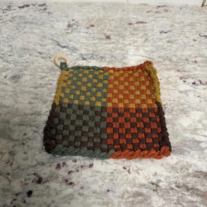 May include: A square, woven hot pad with a wooden ring for hanging. The pad is divided into four sections of colour: green, yellow, orange, and brown. The woven design is a checkerboard pattern.