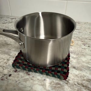 May include: A stainless steel saucepan with a long handle sits on a woven trivet. The trivet has a checkered pattern in red, green, and black. The pan is on a granite countertop.