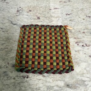 May include: A square, woven hot pad in a checkerboard pattern of green, orange, and burgundy. The edges are trimmed with a braided border in green and burgundy. A small wooden loop is attached for hanging.