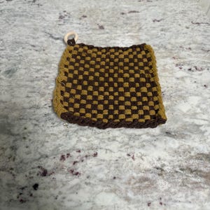 May include: A square, woven potholder in shades of brown and gold. The checkerboard pattern is accented with a darker brown border and a wooden ring for hanging. This kitchen accessory is designed for heat resistance.