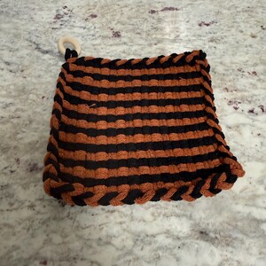 May include: A square, handmade pot holder with alternating horizontal stripes of black and orange. The edges are trimmed with a braided black border. A small, round wooden ring is attached to one corner.