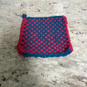 May include: A square, woven hot pad in a checkerboard pattern of teal and magenta. The edges are trimmed with a teal rope-like border. A small wooden ring is attached to one corner for hanging.