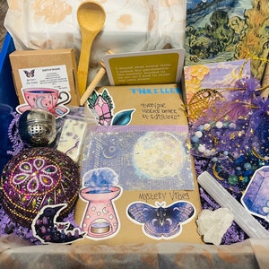 May include: A blue box filled with various items, including a wooden spoon, tea, stickers, a tea infuser, a decorative ball, and small bags. The box contains a variety of purple and blue items.