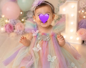 May include: A baby wearing a pastel rainbow-colored tulle dress with butterfly accents. The dress has a rhinestone "ONE" embellishment. The background includes balloons, a lighted number one, and a rainbow.