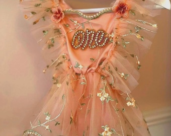 May include: A peach-colored baby dress with floral embroidery and the word "one" embellished with rhinestones. The dress features ruffled sleeves and a tulle skirt, hanging on a white hanger.